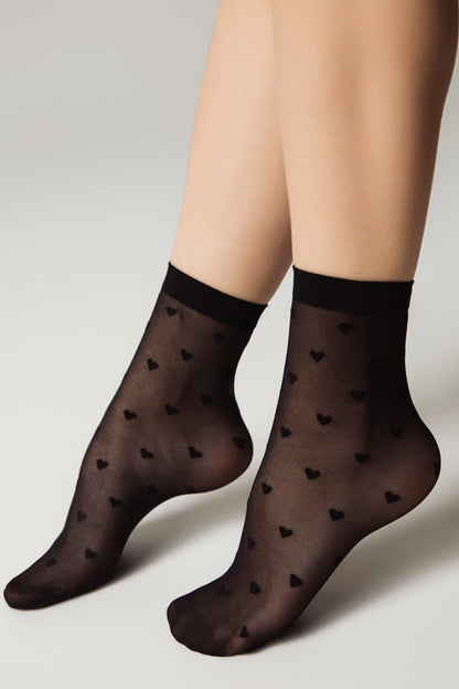 Sheer black ankle socks patterned with small black hearts worn on a pair of legs against a plain light background.