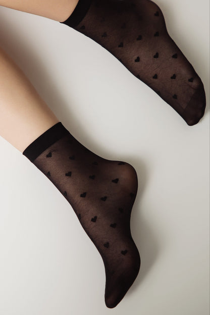 A pair of sheer black ankle socks with a repeating small heart pattern worn on a person's feet against a plain white background.
