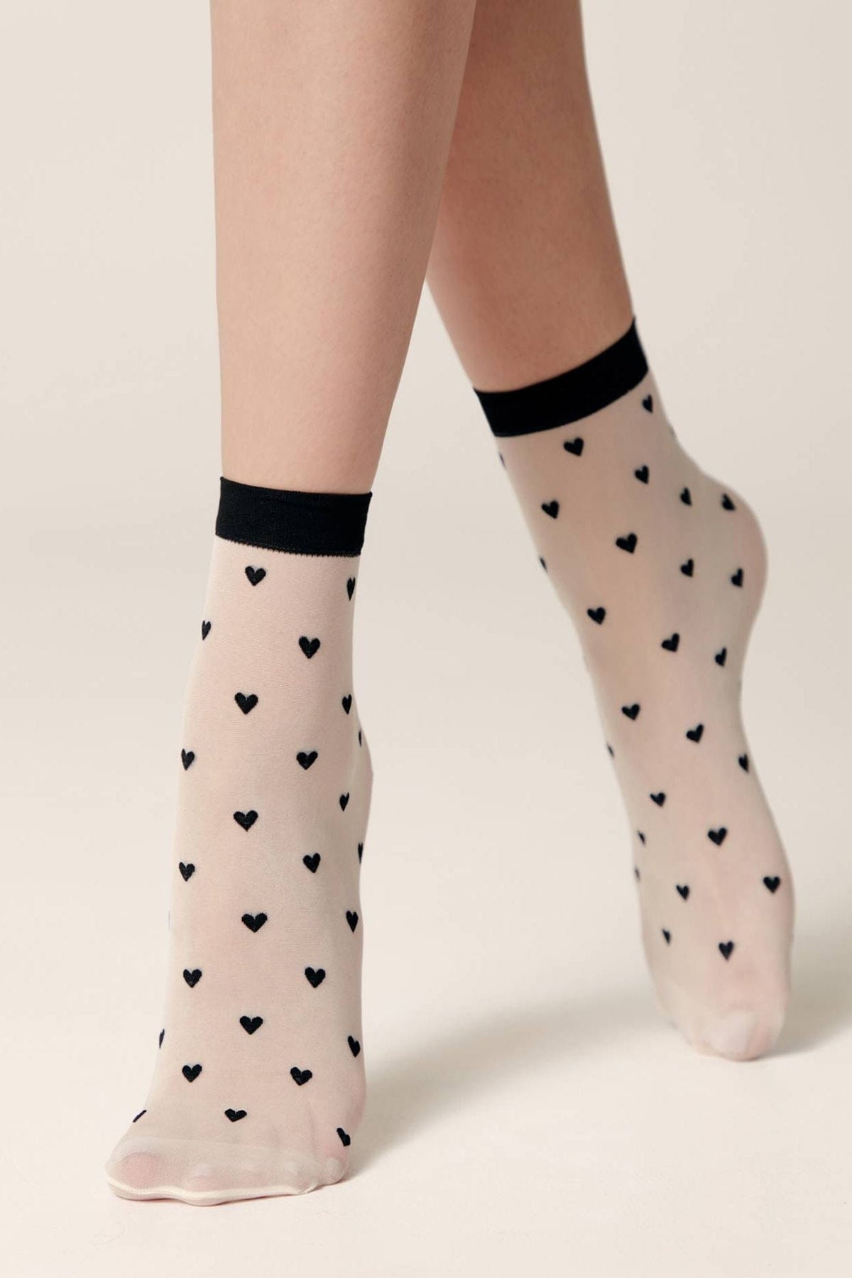 Sheer ankle socks in a light beige tone, featuring an all-over pattern of small black hearts and finished with a solid black trim at the cuffs.