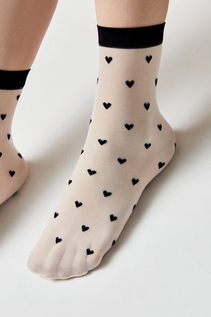 Sheer ankle-length socks in a beige tone, featuring a repetitive pattern of small black hearts and a contrasting black cuff.