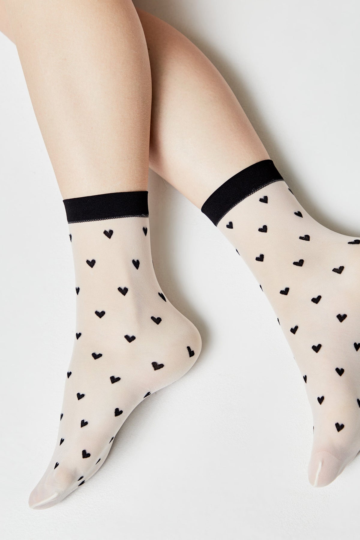 Sheer white ankle socks featuring a black heart pattern and a black ribbed trim worn on a person's feet against a plain white background.