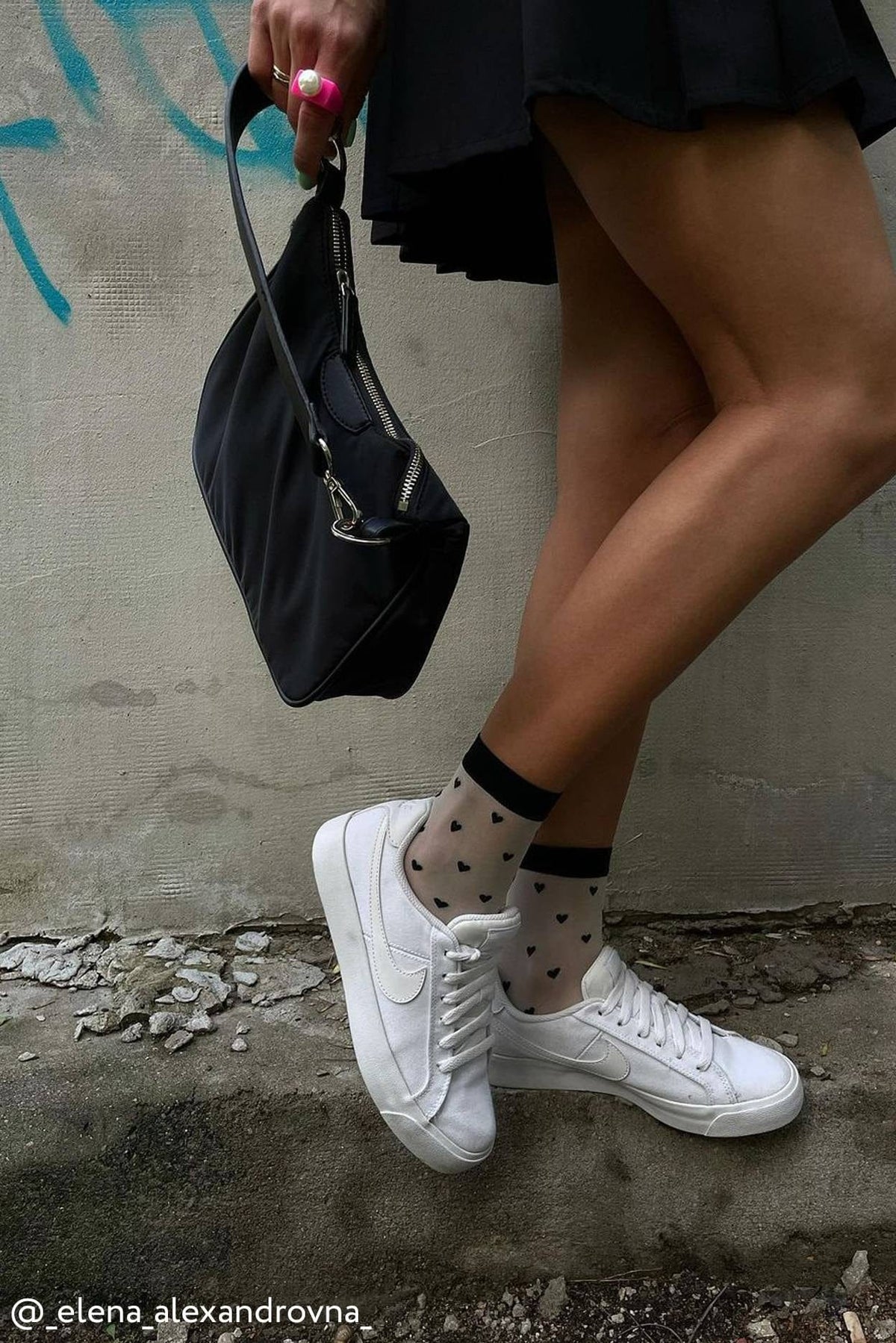 White sneakers with white laces and a side logo swoosh are worn with sheer white socks featuring a black heart pattern and black cuffs.