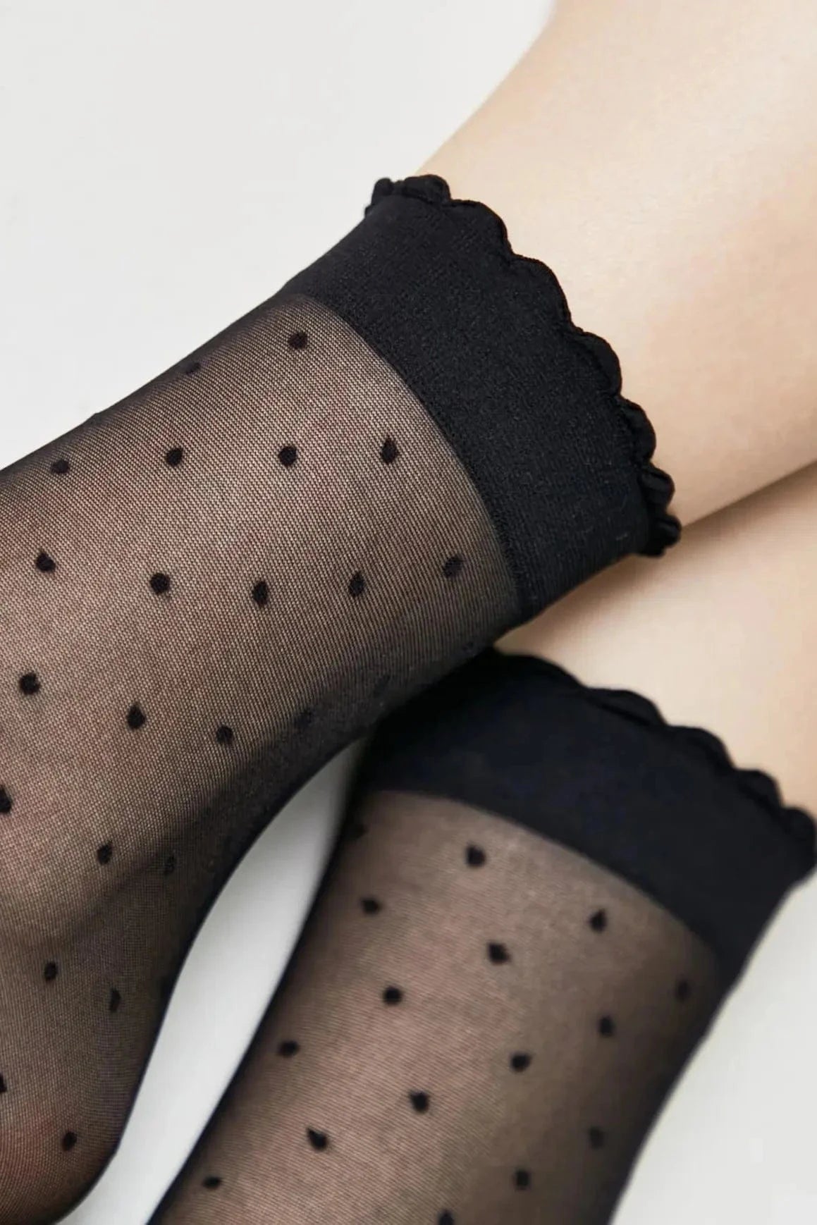 Sheer black ankle socks featuring a black polka dot pattern and a scalloped ruffled edge at the cuff.