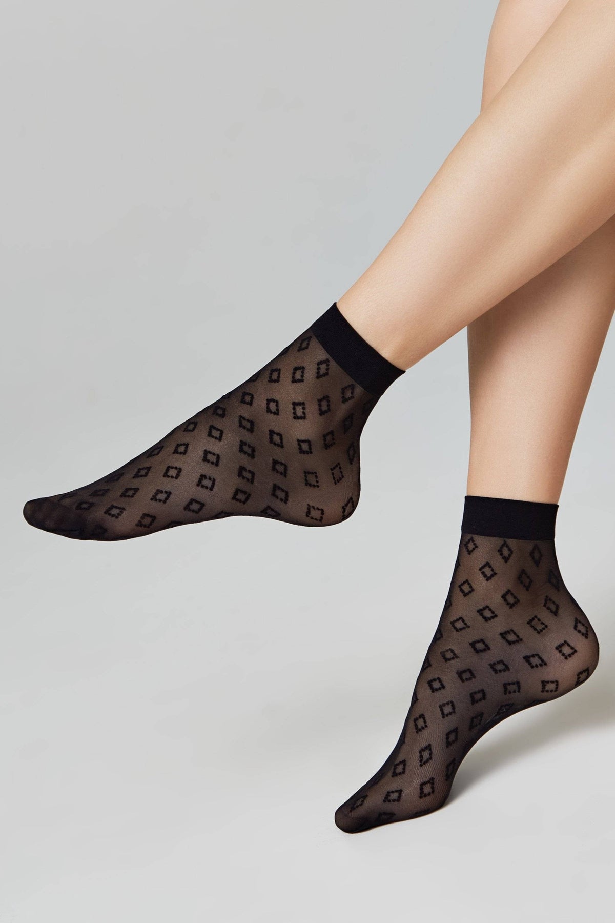 A pair of sheer black ankle socks with a repeating square pattern, worn on a model's feet against a light grey background.