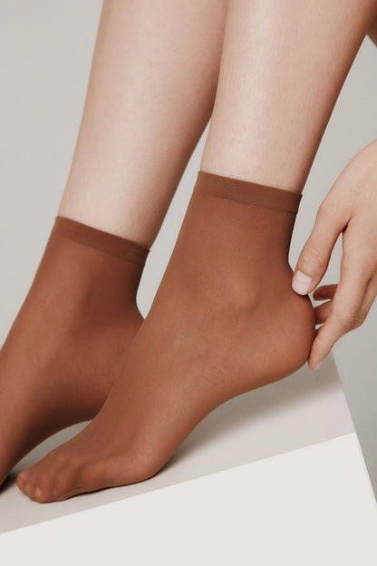 Sheer brown ankle socks worn on feet with a hand lightly touching the heel while resting on a white surface.