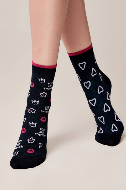 A pair of navy blue crew socks featuring a pattern of white hearts on the right foot and a mix of white crowns, flowers, pink lips, and the phrase YOU ARE ENOUGH on the left foot.