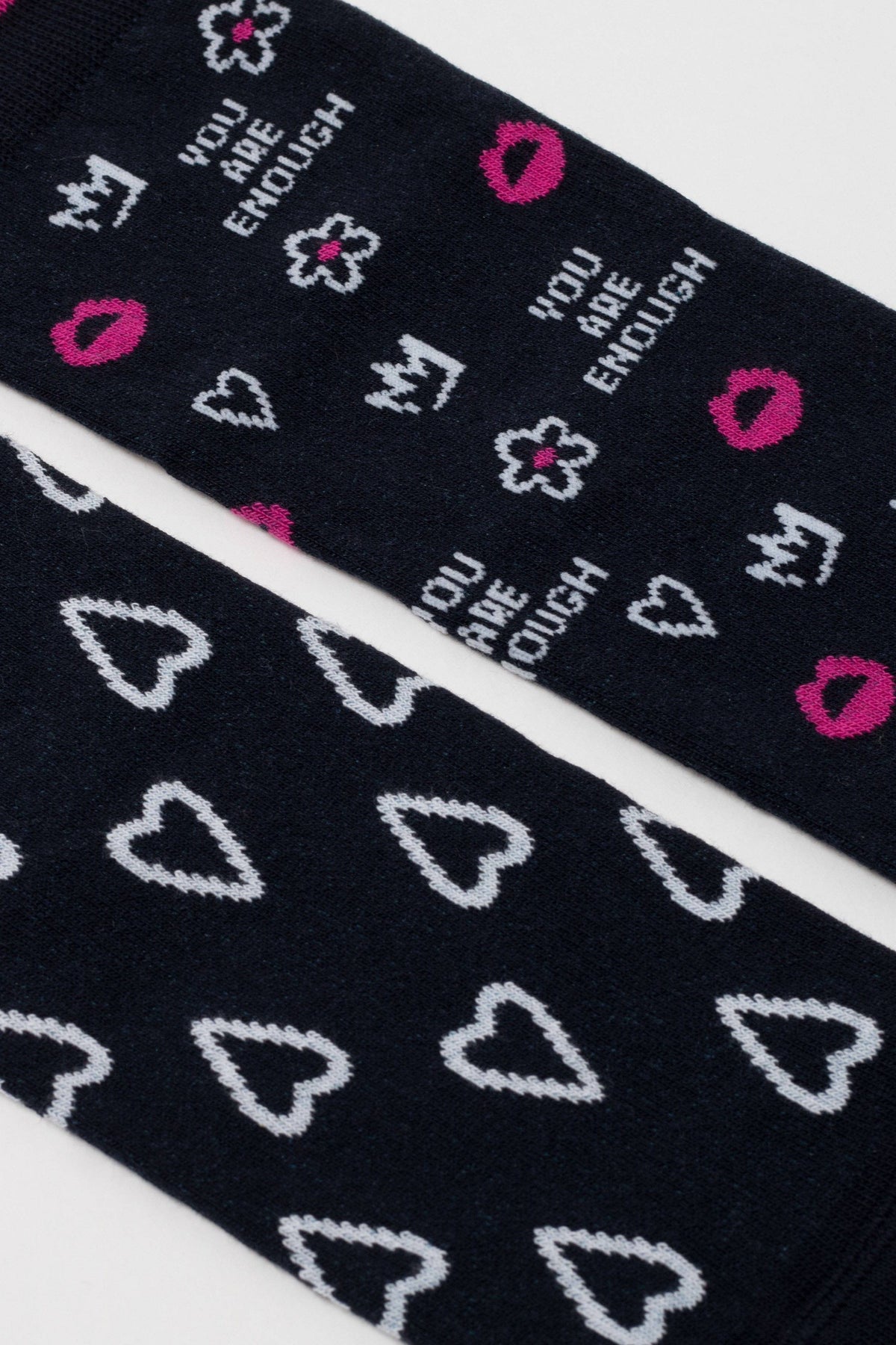 A pair of dark navy blue socks featuring a repeating pattern of white hearts on one sock and a mix of white hearts, crowns, flowers, and the phrase YOU ARE ENOUGH along with small pink lips on the other.
