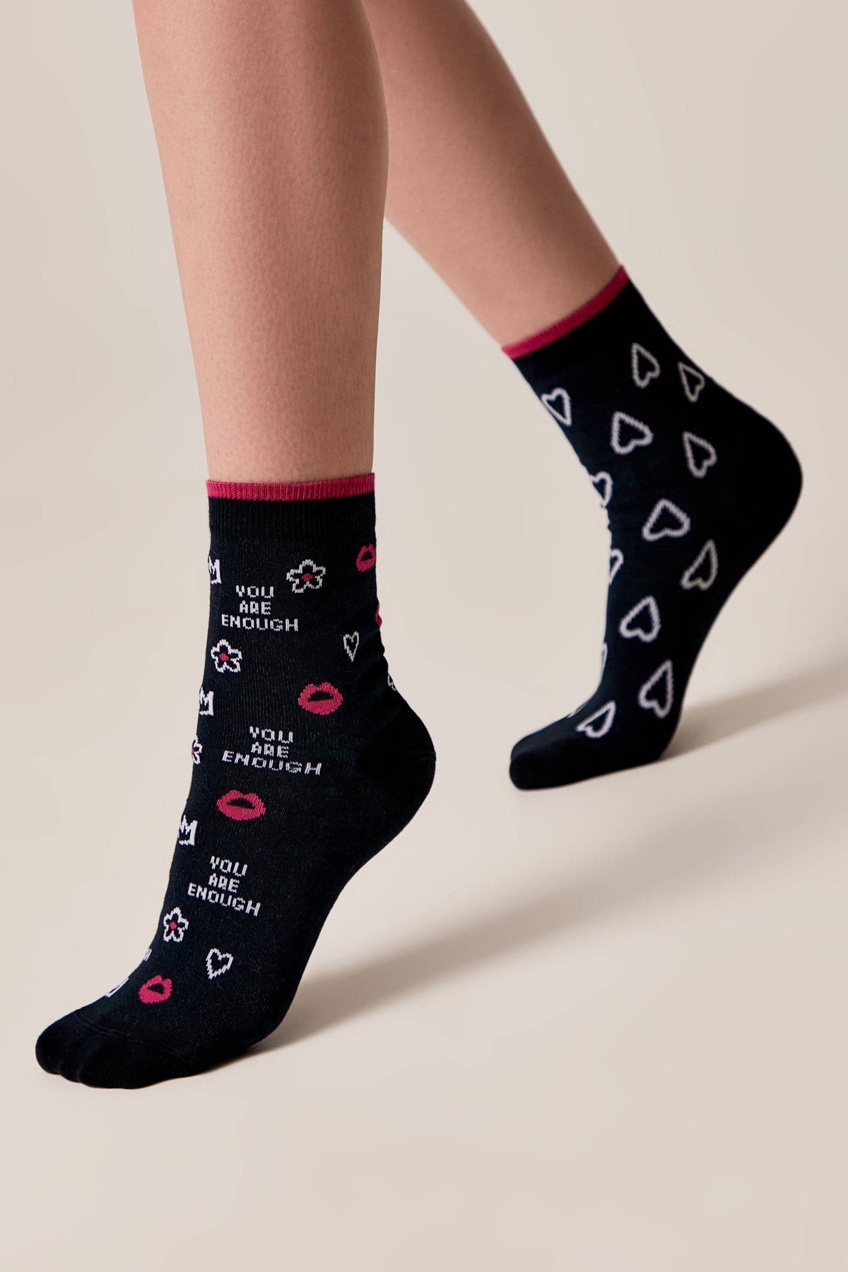 A pair of dark-colored socks featuring an all-over pattern of white hearts, small flowers, pink lips, and the repeated text "YOU ARE ENOUGH" worn on a model's legs against a neutral background.