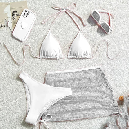 A white three-piece bikini set includes a triangle top with pink trim, matching high-cut bottoms, and a sheer silver mesh skirt, displayed on a textured white surface with a phone case and sunglasses nearby.