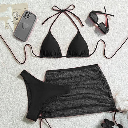 A black bikini set consisting of a triangle top and high-waisted bottom, paired with a matching sheer black mesh mini skirt, sunglasses, and a grey phone case against a white textured background.