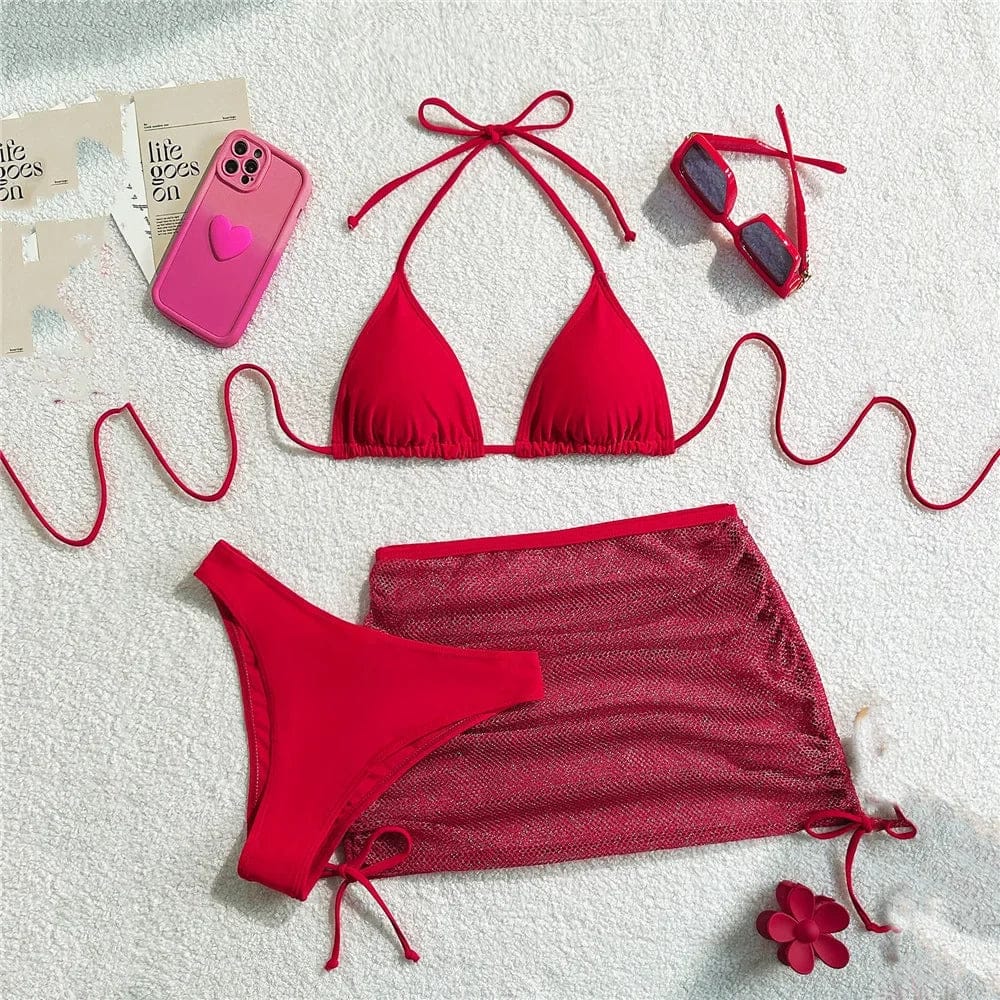 A red bikini set consisting of a triangle top, a matching bottom, and a drawstring mini skirt, arranged on a white textured background with a pink phone case, red sunglasses, and a flower accessory.