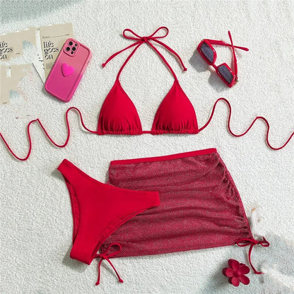 A red bikini set consisting of a triangle top, a matching bottom, and a drawstring mini skirt, arranged on a white textured background with a pink phone case, red sunglasses, and a flower accessory.