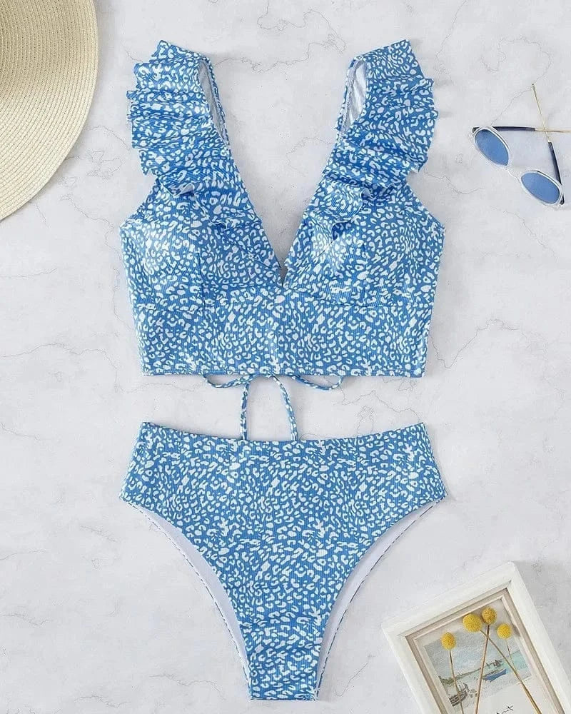 A two-piece bikini set featuring a blue and white leopard print, consisting of a ruffled v-neck crop top and matching high-waisted bottoms, displayed on a marble surface with a hat, sunglasses, and a small picture frame.