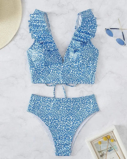 A two-piece bikini set featuring a blue and white leopard print, consisting of a ruffled v-neck crop top and matching high-waisted bottoms, displayed on a marble surface with a hat, sunglasses, and a small picture frame.