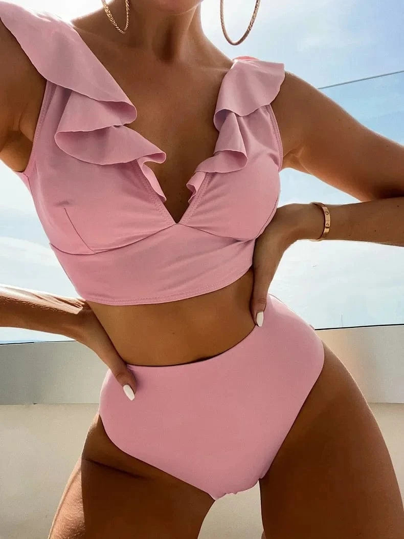 A light pink two-piece bikini featuring a ruffled V-neck crop top and high-waisted bottoms, modeled outdoors against a clear sky.