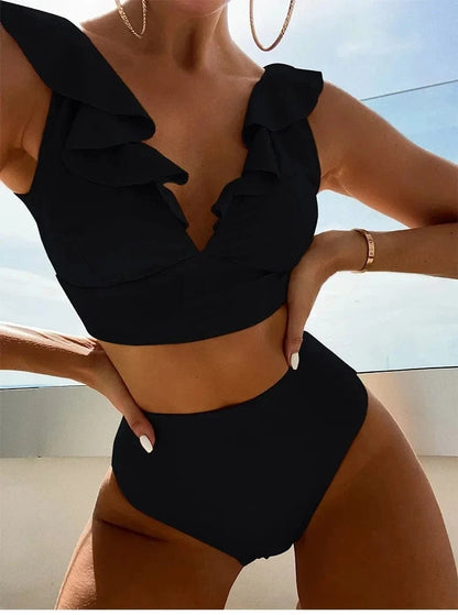 A black two-piece high-waisted bikini with ruffled detailing along the V-neckline worn by a woman posing on a balcony.