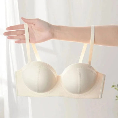 A beige strapless-style bra held up by a person's hand against a soft white background.