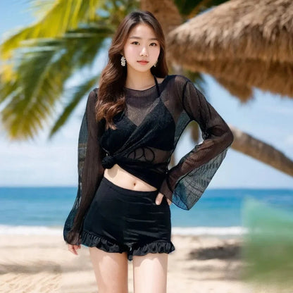 A woman stands on a sandy beach wearing a black two-piece swimsuit with a long-sleeved, semi-sheer black cover-up top and matching high-waisted shorts with a ruffled hem.