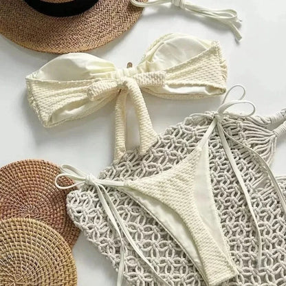 A white two-piece bikini featuring a bandeau top with a central bow and matching tie-side bottoms, laid flat on a decorative macrame mat next to a straw hat.