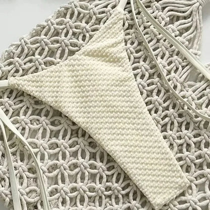 A cream-colored, textured bikini bottom with thin side straps rests on a decorative white macrame background.