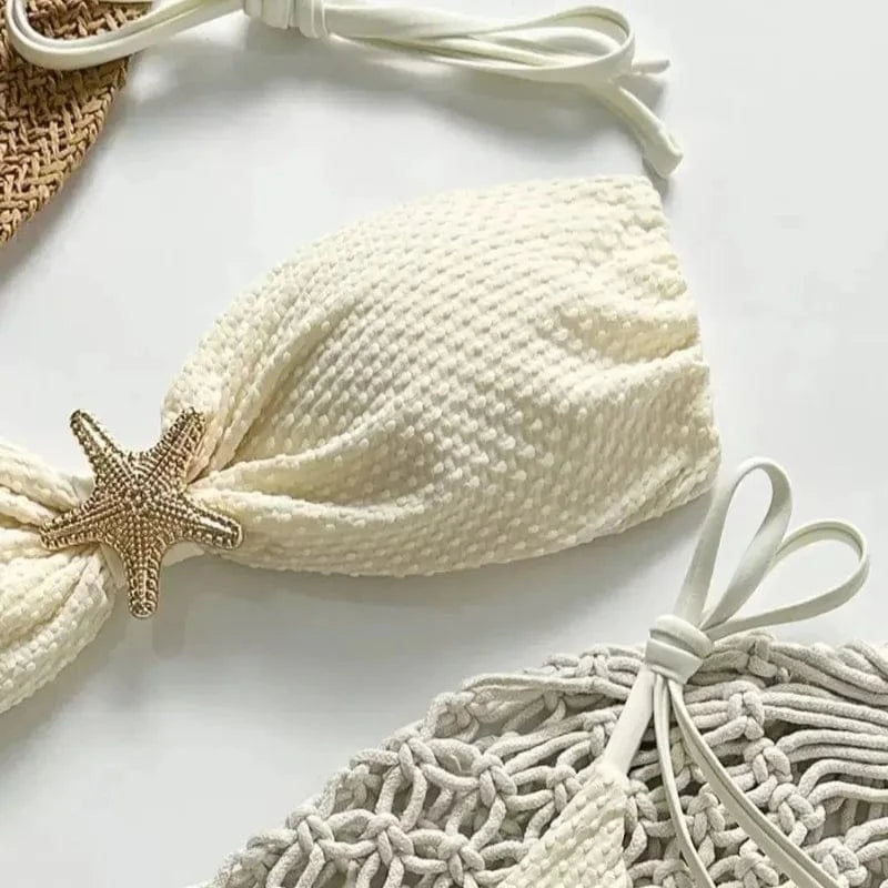 A white textured bikini top featuring a gold-toned starfish embellishment at the center, positioned next to a woven beige material accessory against a white background.