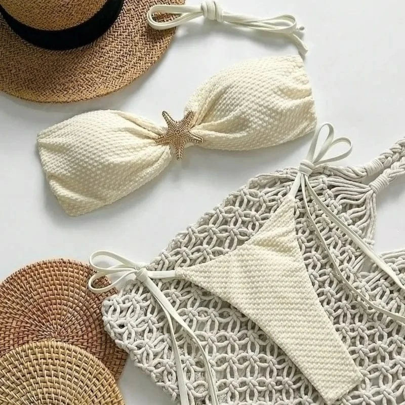 A white two-piece swimsuit with a textured fabric featuring a gold starfish accent on the bandeau top, laid out on a white surface next to a straw hat and a macrame tote bag.