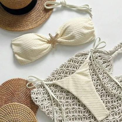 A white two-piece swimsuit with a textured fabric featuring a gold starfish accent on the bandeau top, laid out on a white surface next to a straw hat and a macrame tote bag.