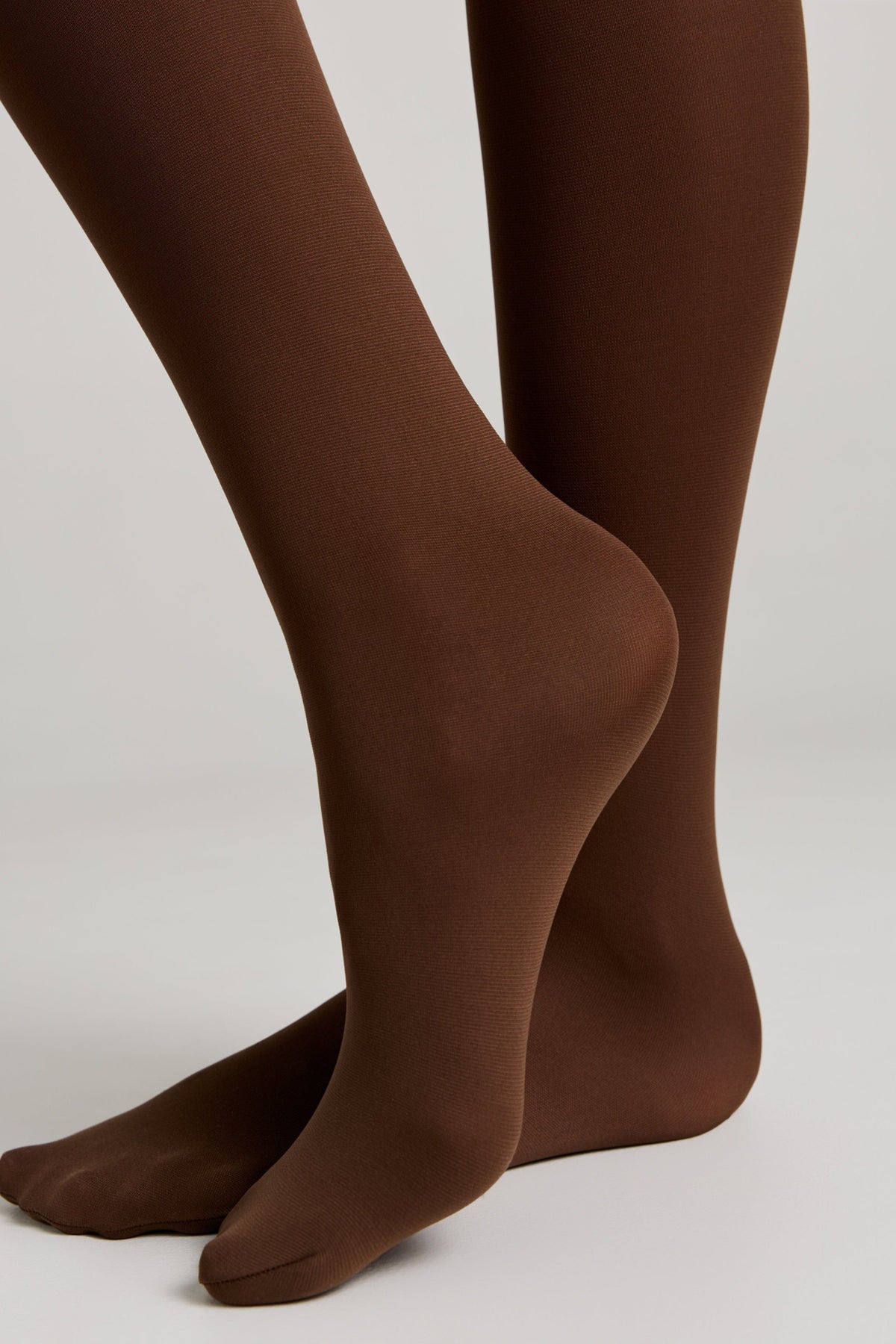 A pair of opaque brown tights worn on the lower legs and feet against a plain light gray background.