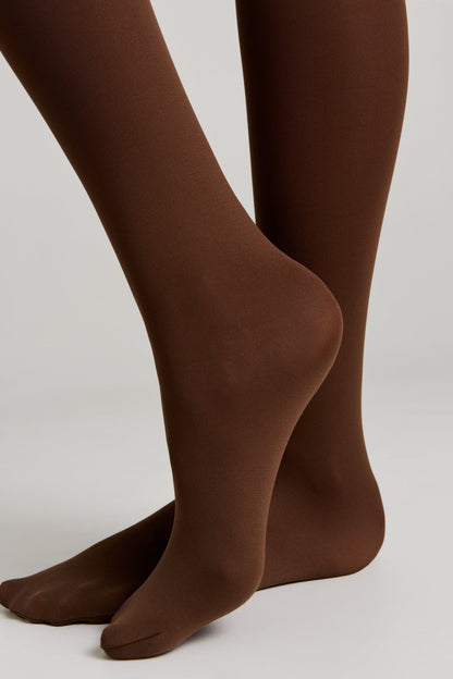 A pair of opaque brown tights worn on the lower legs and feet against a plain light gray background.