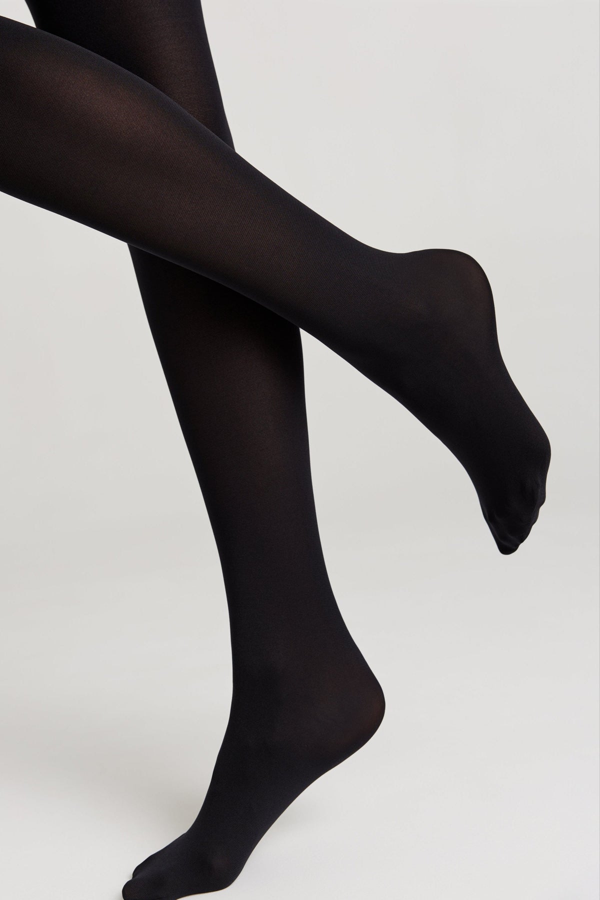 Opaque black tights worn on a person with legs crossed against a plain light gray background.