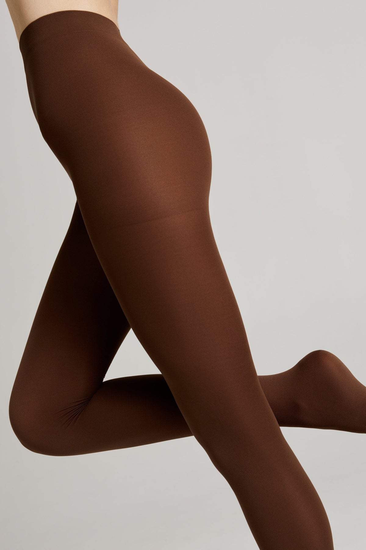A pair of opaque chocolate brown tights modeled on a person's legs against a plain light gray background.