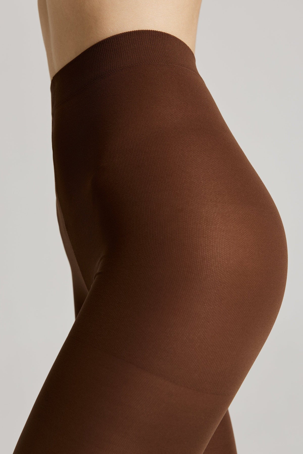 A close-up side view of a person wearing opaque dark brown tights with a high-waisted fit against a plain light gray background.