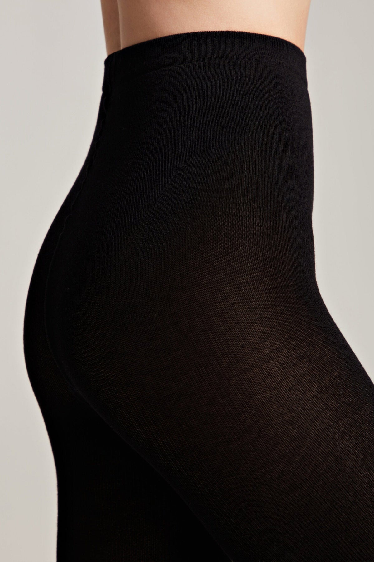 A close-up side view of a person wearing black opaque tights on a plain light grey background.