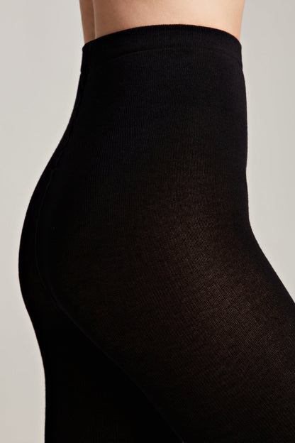 A close-up side view of a person wearing black opaque tights on a plain light grey background.