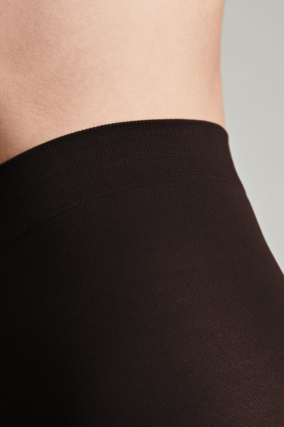 A close-up view of a pair of solid dark brown opaque tights, highlighting the textured knit fabric and the wide elasticized waistband against skin.