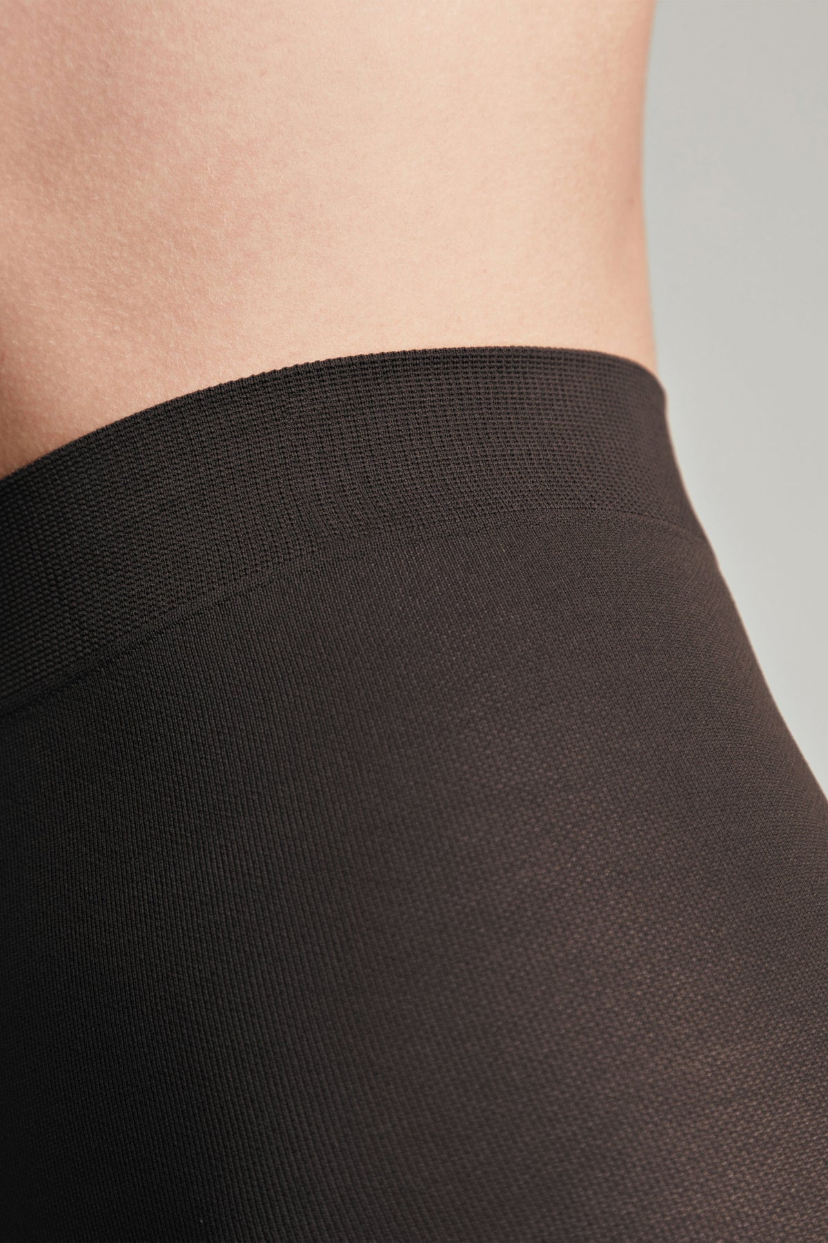 Close-up of a dark brown elastic waistband on smooth opaque tights worn against light skin.