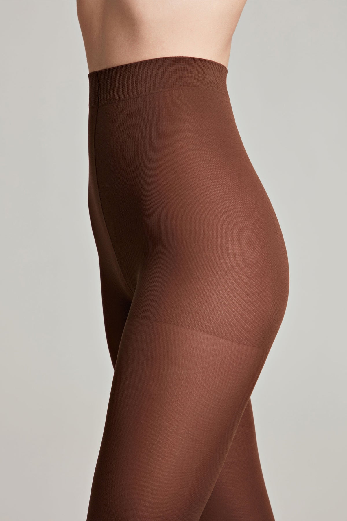 A close-up side profile of a person wearing opaque brown tights with a high-waisted control top against a plain light gray background.