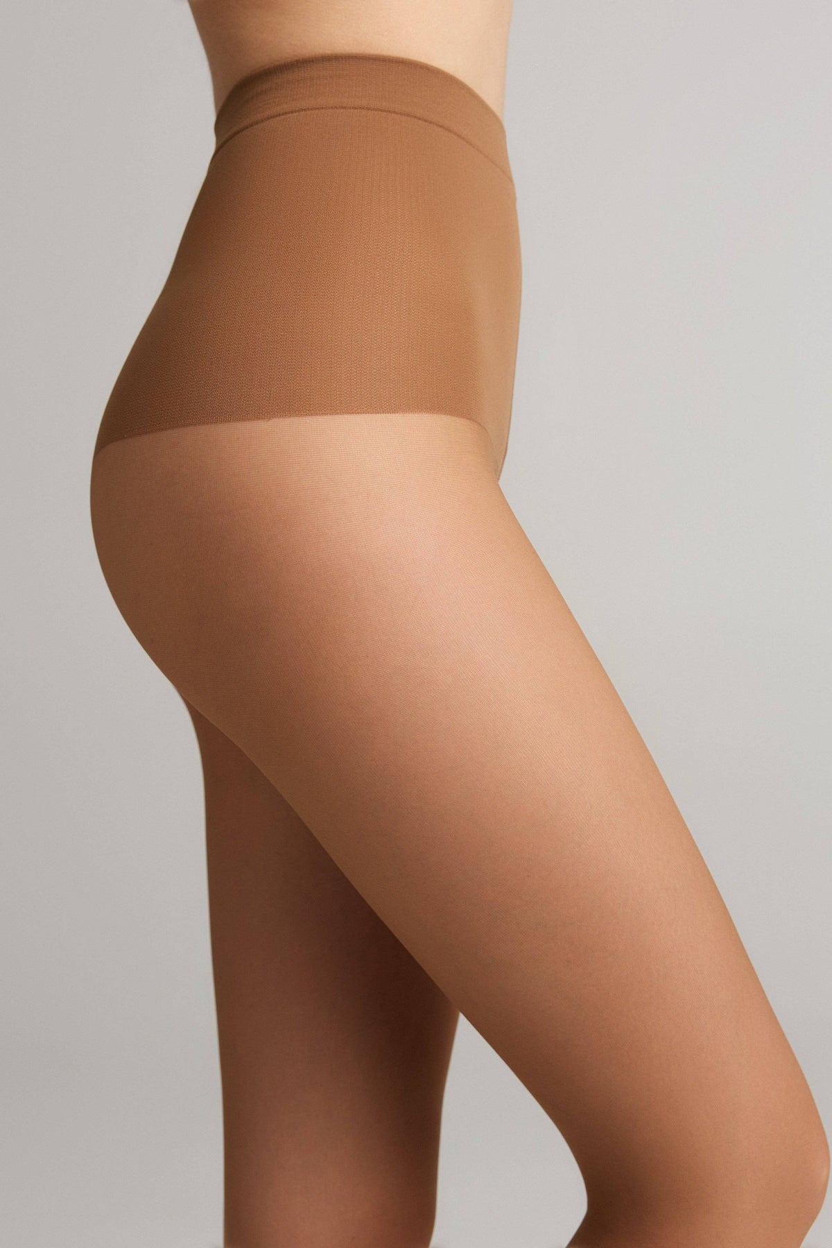 A close-up side view of a person wearing sheer tan hosiery that features a darker, opaque high-waisted control top against a plain light gray background.