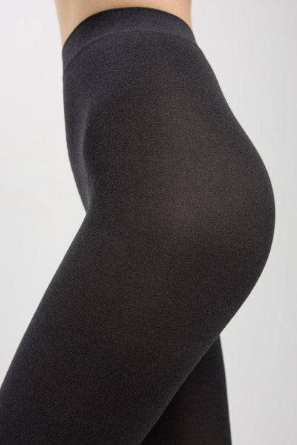 A close-up side view of a person wearing opaque, form-fitting black leggings against a plain light gray background.