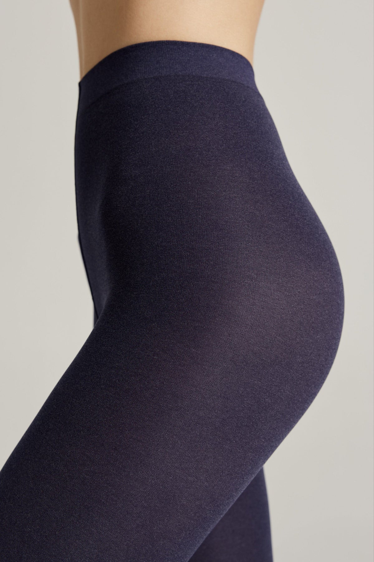 A close-up side view of a person wearing high-waisted, solid dark navy blue opaque leggings against a plain light grey background.