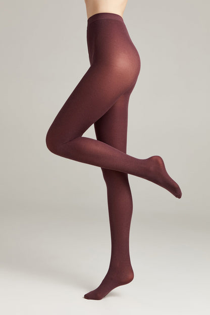 A person stands on one foot wearing opaque, deep burgundy-colored tights against a solid neutral background.