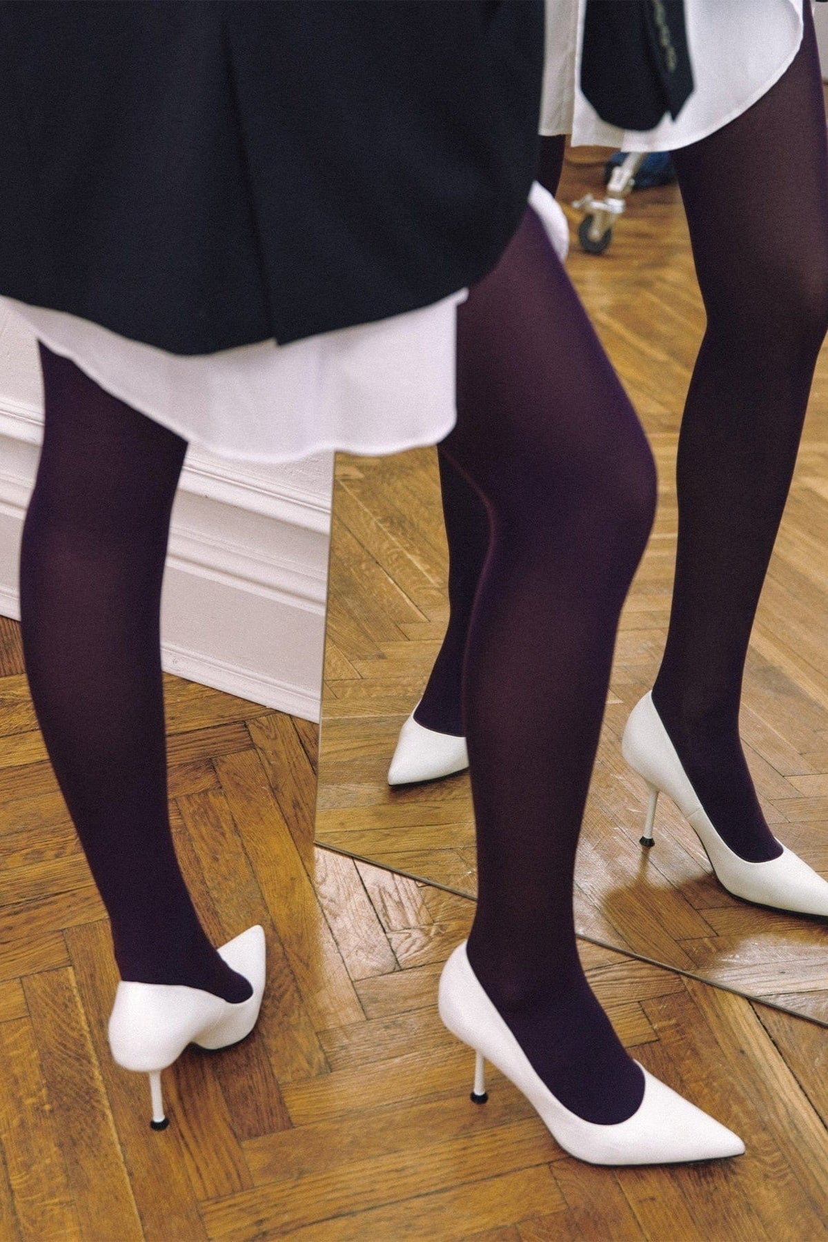 White high-heeled pointed-toe pumps worn with opaque dark purple tights standing on a wooden herringbone floor in front of a mirror.