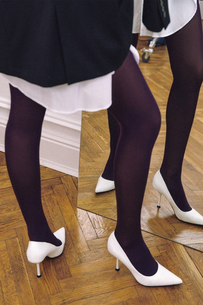 White high-heeled pointed-toe pumps worn with opaque dark purple tights standing on a wooden herringbone floor in front of a mirror.