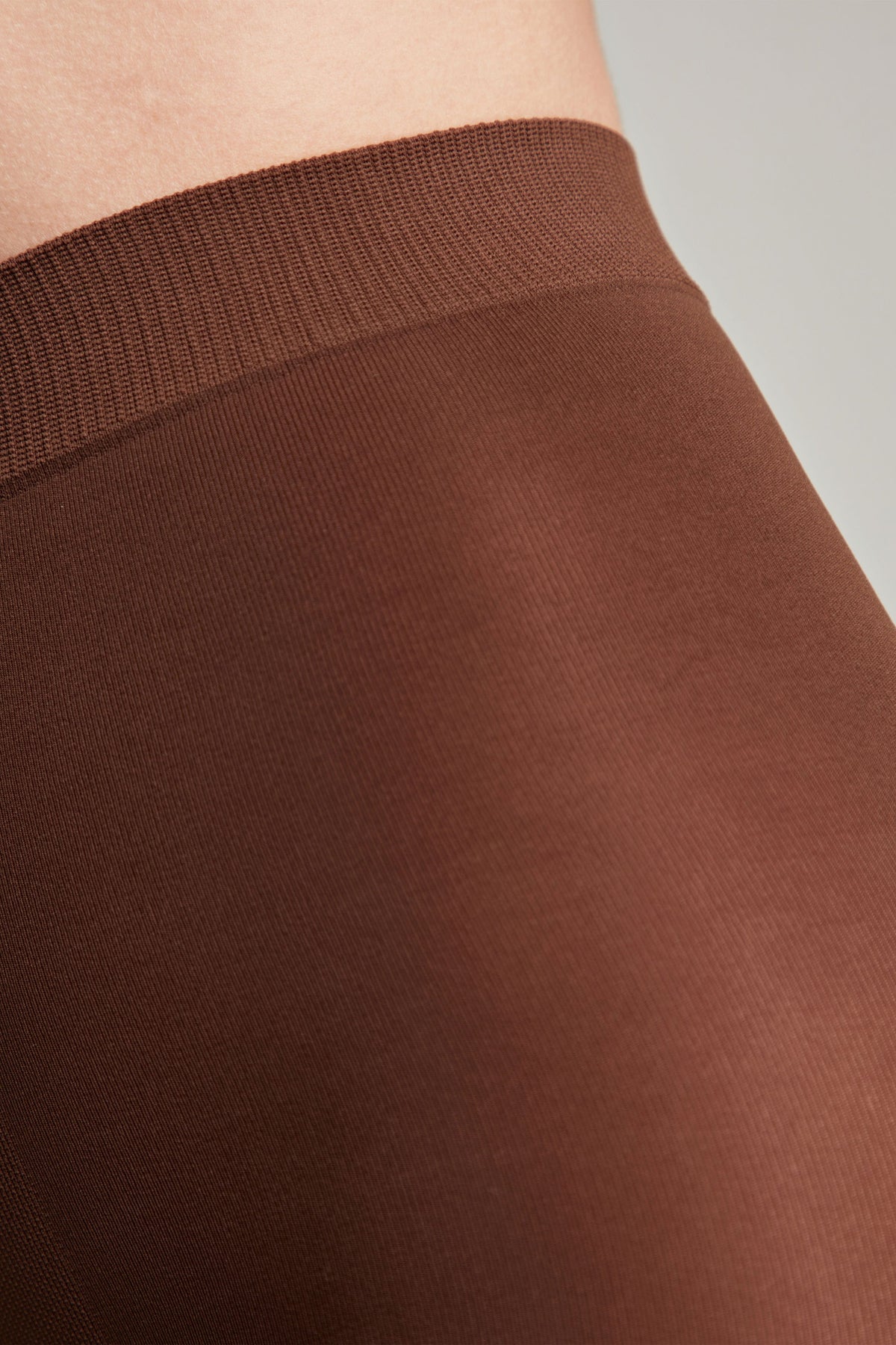A close-up of brown tights featuring a ribbed elastic waistband on a person's upper thigh.