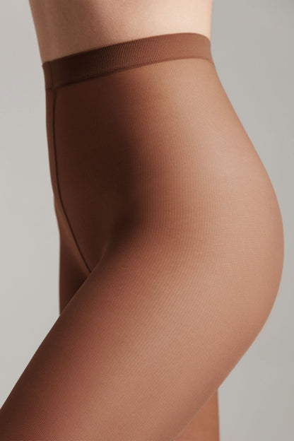 A close-up view of the side profile of a person wearing sheer brown tights with a high waistband against a plain gray background.