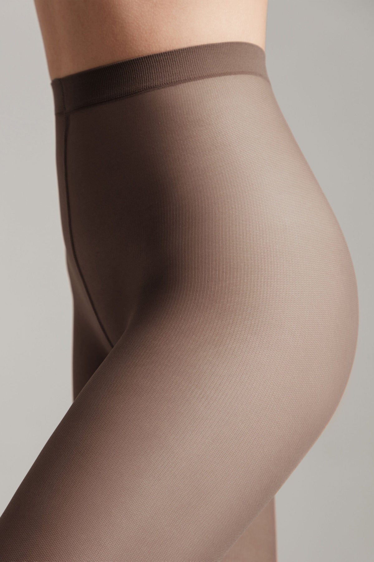 A close-up view of a person wearing sheer, mocha-colored tights that feature a seamless design and an elastic waistband.