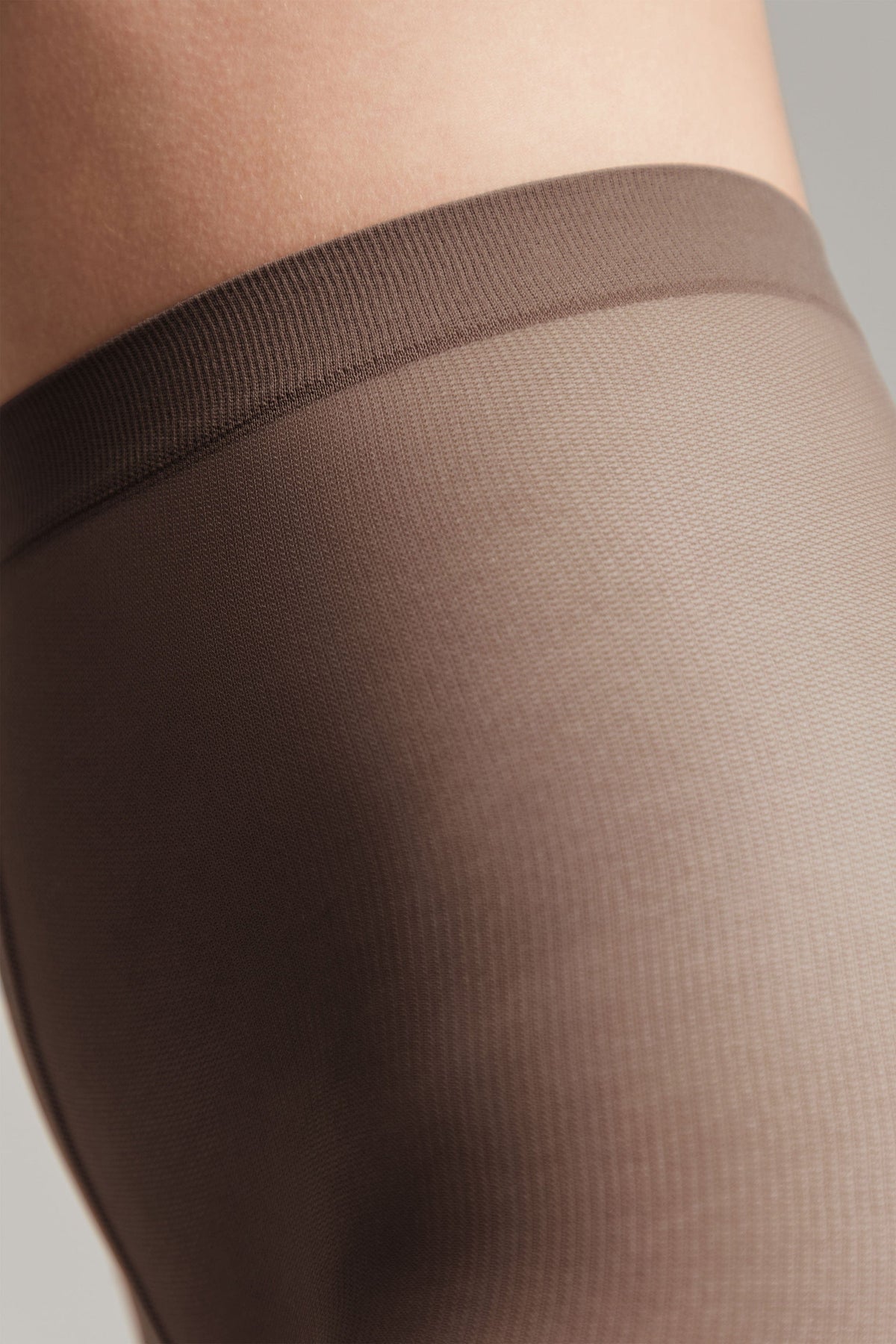 Sheer brown pantyhose worn on a person's upper leg, featuring a smooth, knitted texture and a wide, reinforced waistband.