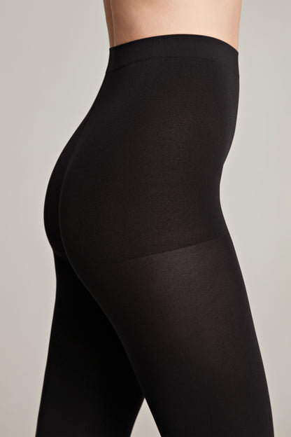 Black opaque tights worn on a person, featuring a high-waisted design that reaches the mid-torso against a neutral light grey background.