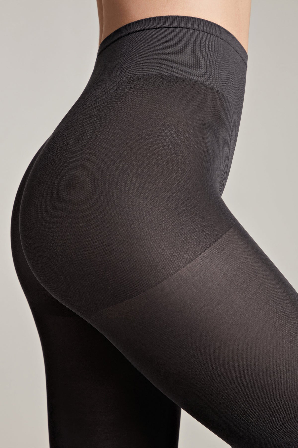 A close-up side view of a person wearing black opaque high-waisted tights against a plain light gray background.