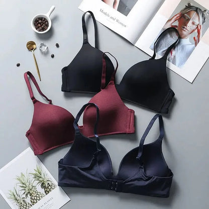 Three bras in black, burgundy, and dark navy blue are arranged on a flat grey surface alongside a small bowl of coffee beans, a gold spoon, and two open books.
